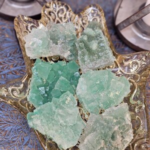 Puede incluir: Cristales de fluorita verde en un plato decorativo dorado en forma de mano. Los cristales, de diferentes tonos de verde, tienen una estructura cristalina. El plato ornamentado con patrones detallados se encuentra sobre una superficie azul.