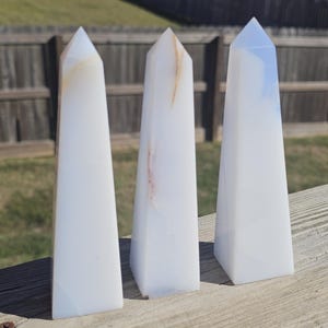 May include: Three white obelisks with subtle color variations and pointed tops are arranged on a wooden surface. The obelisks have a smooth, polished appearance, and are likely made of stone or crystal.