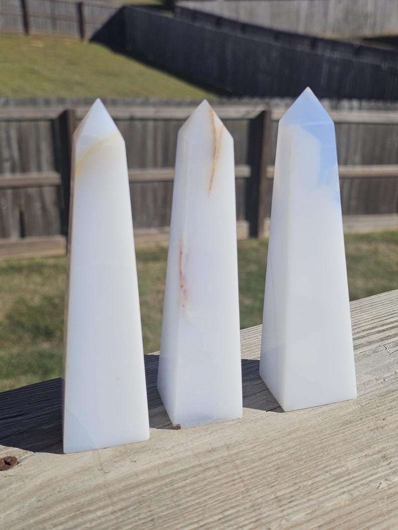 May include: Three white obelisks with subtle color variations and pointed tops are arranged on a wooden surface. The obelisks have a smooth, polished appearance, and are likely made of stone or crystal.
