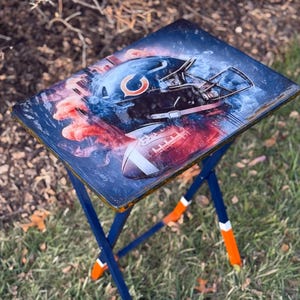 May include: A folding TV tray with a blue frame and a tabletop featuring a Chicago Bears football helmet and football design. The legs have orange and white accents. The background has a smoky effect.