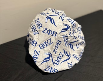 May include: A white satin bonnet with a blue pattern of the letters "ZΦB" repeated across the surface. The bonnet is gathered at the top and appears to be made of a lightweight material. The background is a black surface.