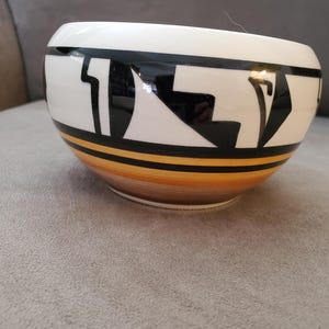 May include: A white ceramic bowl with black and gold geometric designs. The bowl has a rounded shape and a wide rim.