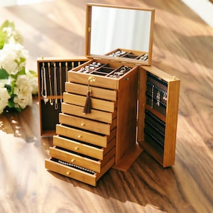 May include: Wooden jewelry box with multiple drawers and compartments. The box is open, revealing necklaces, earrings, and other jewelry. A mirror is attached to the top. The box is made of light brown wood and has a tassel.