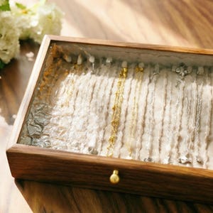 Op de afbeelding: Een rechthoekige houten sieradendoos met een glazen bovenkant, met diverse kettingen. De binnenkant heeft een lichtgekleurde, getextureerde stof. De doos heeft een kleine, ronde, goudkleurige knop. Het hout heeft een warme, bruine tint.