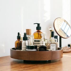 Puede incluir: Una bandeja giratoria de madera oscura exhibe varios productos cosméticos y para el cuidado de la piel. Se ven botellas de vidrio ámbar y transparente, un pequeño espejo y etiquetas. La bandeja está sobre una superficie de madera.