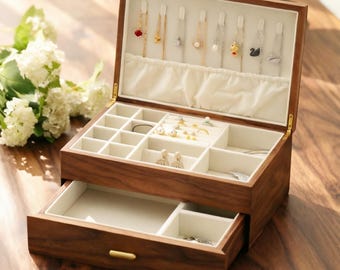 Puede incluir: Una caja de joyería de madera abierta, que revela compartimentos para joyas. La caja es marrón con un interior crema y herrajes dorados. La sección superior tiene espacios para anillos, pendientes y collares.