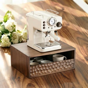Può includere: Una macchina per caffè espresso color crema è posizionata su un supporto rettangolare in legno. Il supporto presenta un cassetto frontale con un motivo a nido d'ape intagliato e testurizzato, contenente diverse tazze in ceramica bianca.