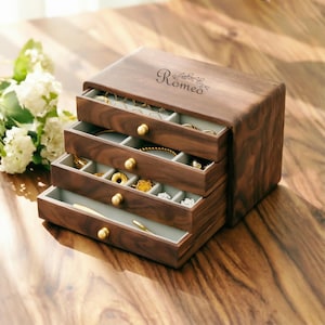 May include: A dark wooden jewelry box with four drawers, each lined with light grey fabric. The box has gold-coloured knobs and the word "Romeo" is engraved on the top. The drawers are open, displaying various pieces of jewellery.