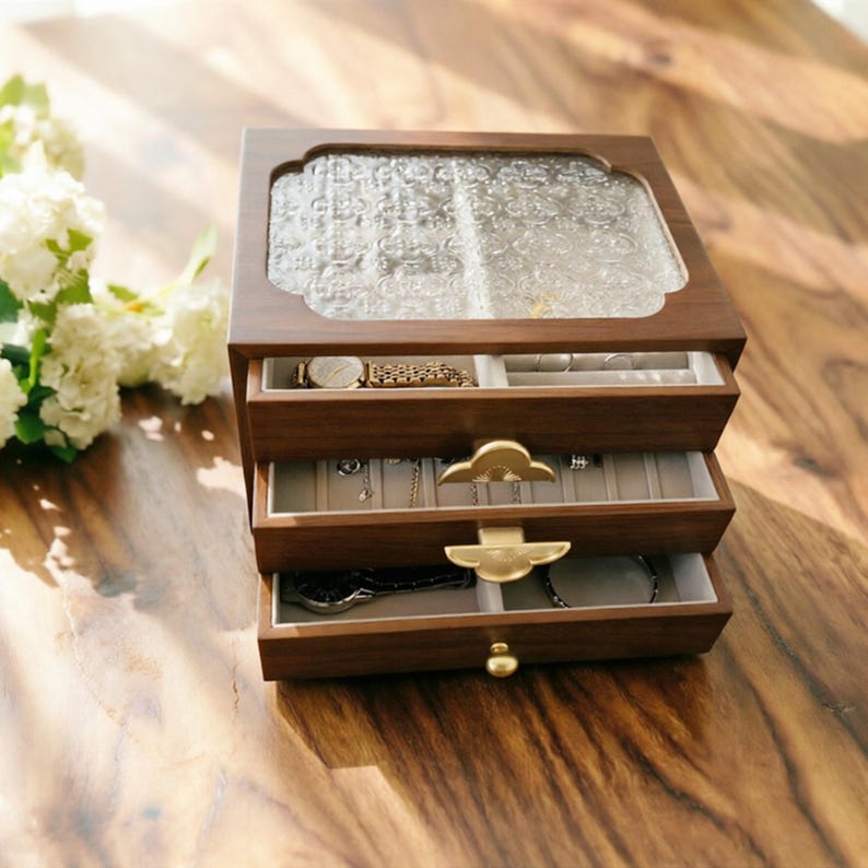 May include: A wooden jewelry box with three drawers and a glass top. The box is filled with jewelry, including a watch, necklaces, and bracelets. The drawers have gold-colored handles. The box is sitting on a wooden surface.
