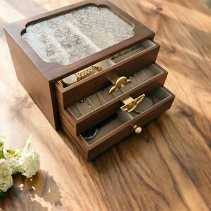 May include: A wooden jewelry box with three drawers and a glass top. The box is made of dark wood and has gold-colored drawer pulls. The drawers are lined with a light-colored fabric and contain jewelry, including a watch and necklaces.