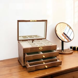 Op de afbeelding: Een houten sieradendoos met een scharnierende spiegel, open om compartimenten en laden te onthullen. De doos heeft een warme bruine kleur met goudkleurige hardware. Een ronde spiegel op een standaard staat rechts.
