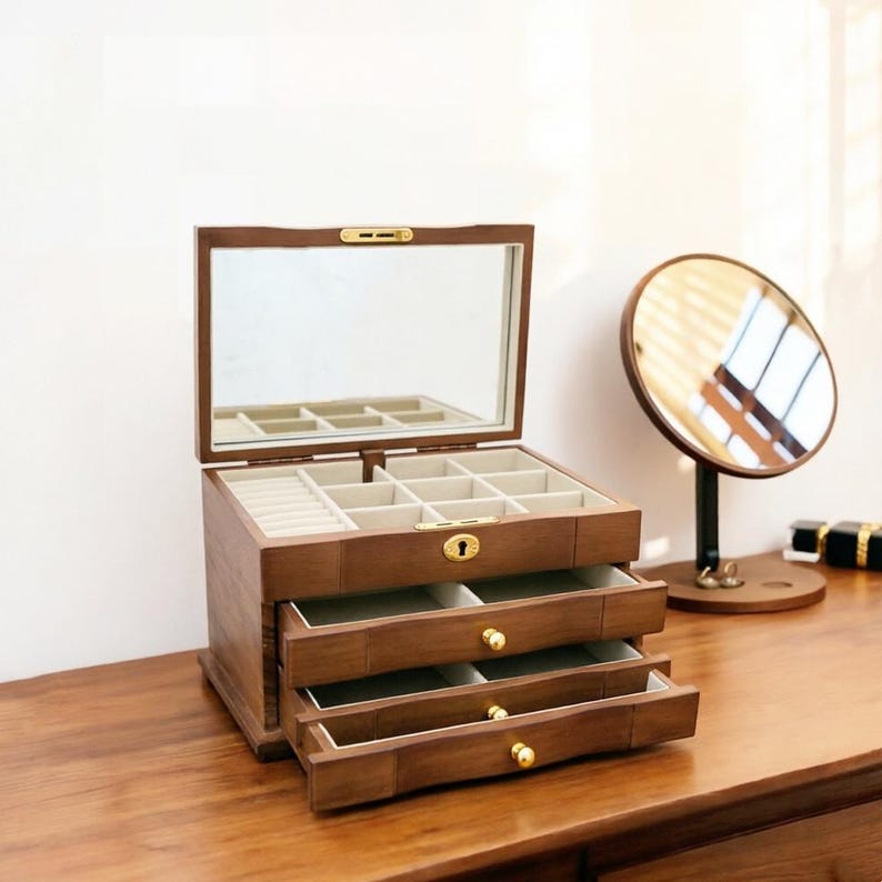 Op de afbeelding: Een houten sieradendoos met een scharnierende spiegel, open om compartimenten en laden te onthullen. De doos heeft een warme bruine kleur met goudkleurige hardware. Een ronde spiegel op een standaard staat rechts.