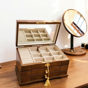 Op de afbeelding: Een houten sieradendoos met een spiegeldeksel, open om meerdere compartimenten met beige stof te onthullen. De doos heeft een goudkleurige sluiting en een decoratieve kwast. Een ronde spiegel op een standaard is zichtbaar op de achtergrond.