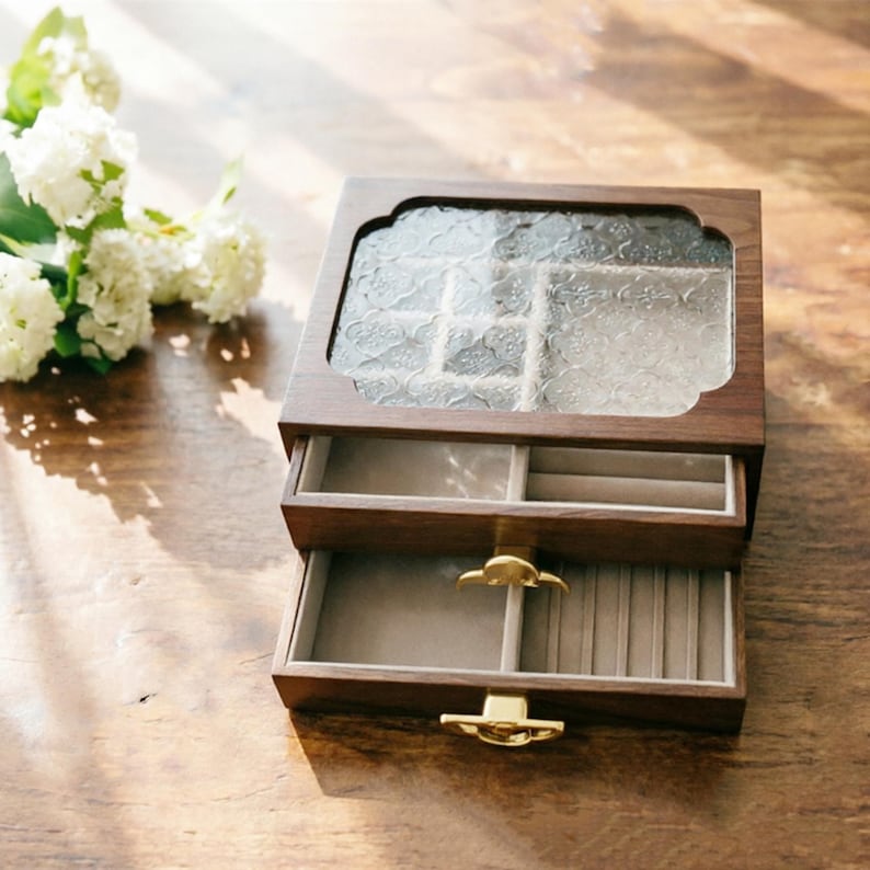 May include: A wooden jewelry box with two drawers and a glass top. The box is made of dark wood and has a decorative glass panel on the top. The drawers are lined with a light-colored fabric and have gold-colored handles.