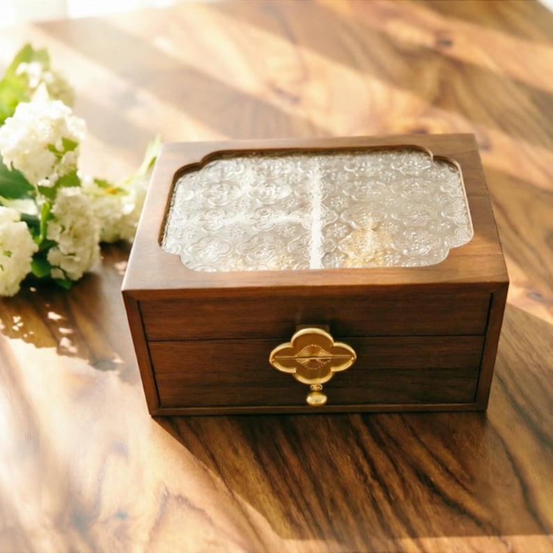 May include: A wooden jewelry box with a clear, patterned glass top and a gold-colored, four-leaf clover shaped handle. The box is a rich brown color and sits on a wooden surface.