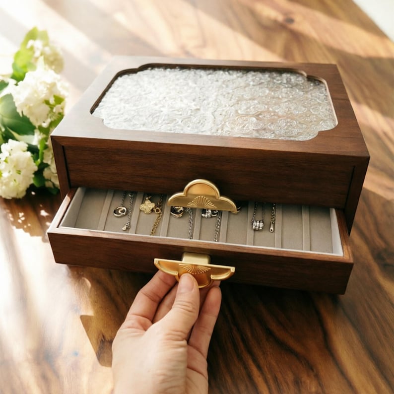 May include: A wooden jewelry box with a clear, textured glass top and a drawer. The drawer is open, revealing several necklaces hanging inside. The box has gold-colored hardware and is sitting on a wooden surface.