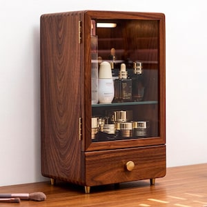 Op de afbeelding: Een donkerbruine houten make-upkast met een glazen deur en een lade. De kast heeft een goudkleurige knop en pootjes. De kast is gevuld met diverse cosmetische producten. Ongeveer 30 cm breed.