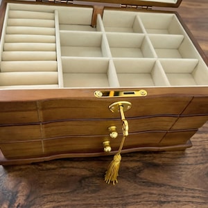 Op de afbeelding: Een houten sieradendoos met meerdere compartimenten en lades. De binnenkant is bekleed met een lichtbeige stof. De doos heeft een sleutel en een gouden kwast. De buitenkant is warmbruin.