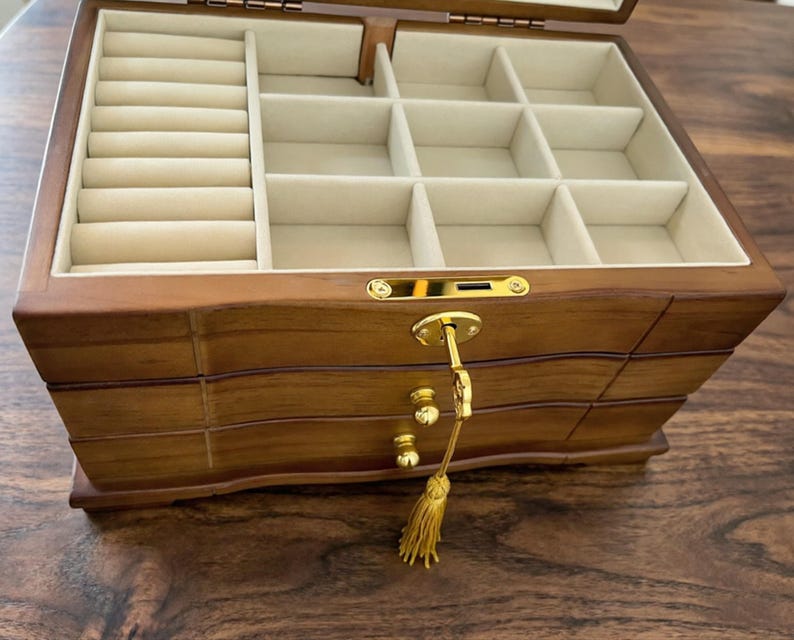 Op de afbeelding: Een houten sieradendoos met meerdere compartimenten en lades. De binnenkant is bekleed met een lichtbeige stof. De doos heeft een sleutel en een gouden kwast. De buitenkant is warmbruin.