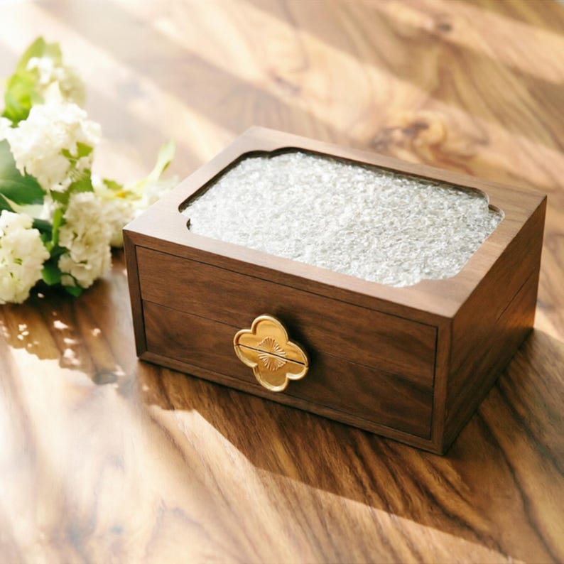 May include: A rectangular wooden box with a glass top and a gold-colored floral-shaped handle. The box is made of dark brown wood and sits on a wooden surface. White flowers are in the background.