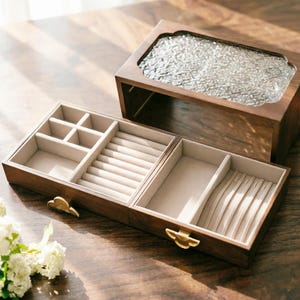 May include: A wooden jewelry box with a clear, textured glass top and a light-colored interior. The box is open, revealing compartments for rings, necklaces, and other jewelry. The box has a warm, brown finish and gold-colored clasps.