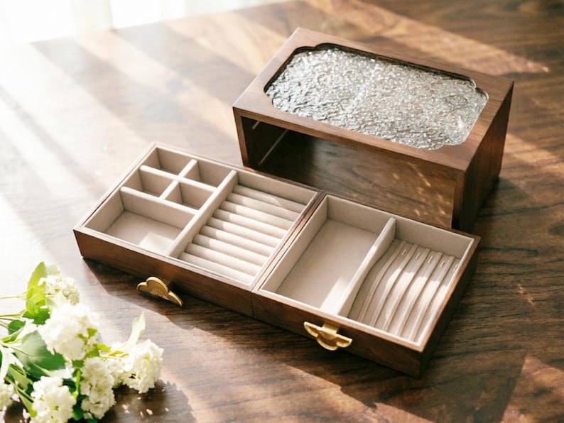 May include: A wooden jewelry box with a clear, textured glass top and a light-colored interior. The box is open, revealing compartments for rings, necklaces, and other jewelry. The box has a warm, brown finish and gold-colored clasps.