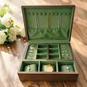 Puede incluir: Caja de joyería de madera abierta con interior de terciopelo verde. La caja contiene anillos, collares, pendientes, un reloj dorado y pulseras. La caja está abierta, revelando múltiples compartimentos para almacenamiento. El exterior es de color marrón oscuro.