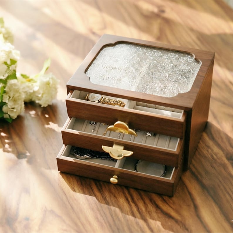 May include: A wooden jewelry box with three drawers and a glass top. The box is a warm brown color and has gold-colored drawer pulls. The drawers are open, revealing jewelry such as a watch, necklaces, and bracelets. The top of the box has a decorative, textured glass panel.