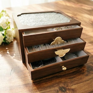 May include: A three-tiered wooden jewelry box with a glass top and gold-colored hardware. The drawers are open, revealing jewelry and accessories. The box is made of dark wood and sits on a wooden surface.