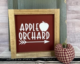 May include: A framed sign with the words "APPLE ORCHARD" in white, along with an apple graphic and an arrow. The sign has a red background and a light brown wooden frame. A decorative fabric apple sits to the right.