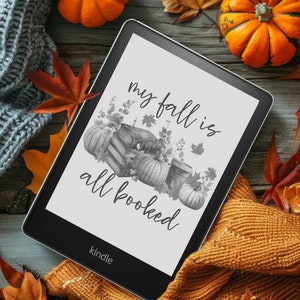 May include: A black and white Kindle e-reader with the text "my fall is all booked" on the screen. The e-reader is surrounded by fall-themed decorations, including pumpkins, leaves, and a knitted blanket.