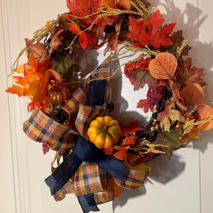 May include: A fall-themed wreath with a plaid bow and two small pumpkins. The wreath is made of brown twigs and has orange, red, and yellow leaves.