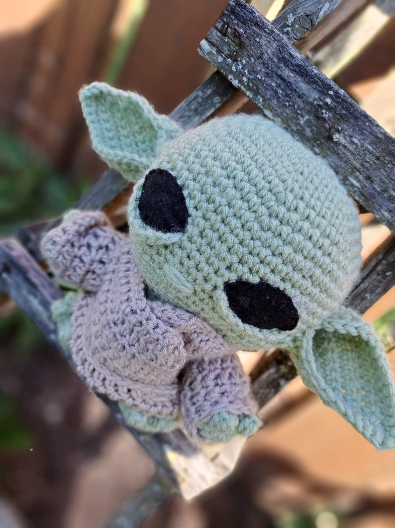 Puede incluir: Un juguete de peluche verde y marr&oacute;n tejido a crochet con orejas grandes y ojos negros. El juguete lleva un su&eacute;ter tejido a crochet marr&oacute;n.