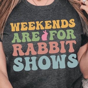 May include: A dark grey heather t-shirt with the text "Weekends are for Rabbit Shows" in colorful letters. The text is in a retro font style.