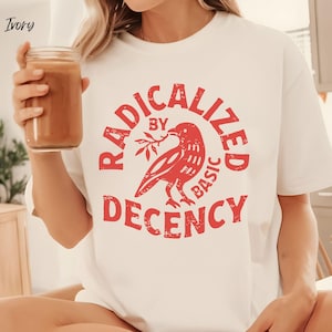 Puede incluir: Camiseta de color marfil con un gráfico rojo que dice "RADICALIZED BY BASIC DECENCY" alrededor de un pájaro que sostiene una ramita. La camiseta de manga corta está hecha de un material suave. Se sostiene un vaso de líquido marrón en la mano.