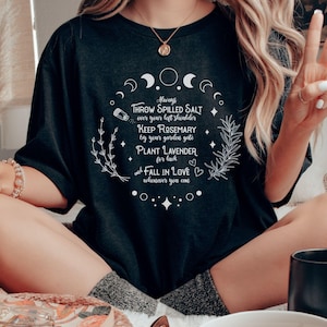 May include: Dark grey t-shirt with a white circular design featuring text about luck and love. The design includes crescent moons, stars, and sprigs of herbs. The text reads "Throw spilled salt over your left shoulder, Keep Rosemary by your garden gate, Plant Lavender for luck, and Fall in Love wherever you can."