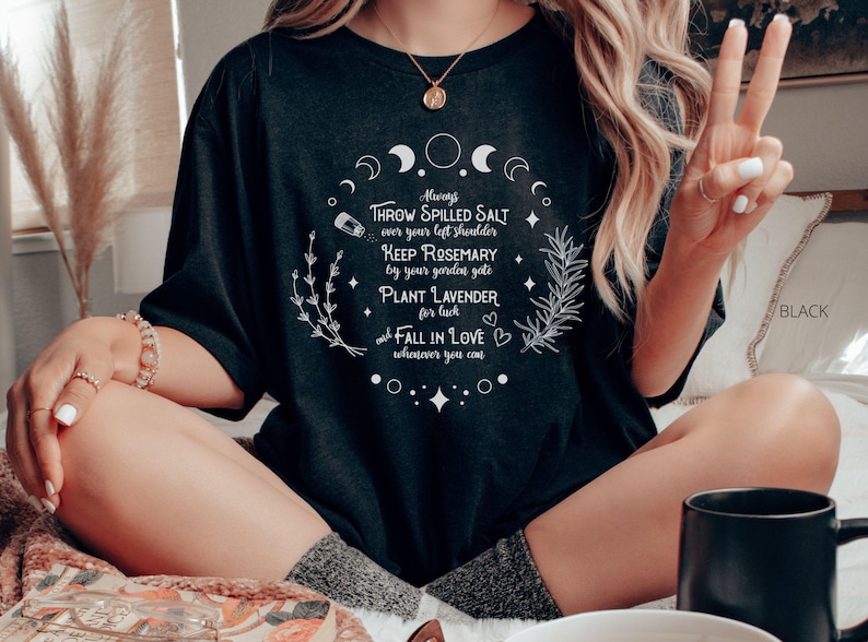May include: Black t-shirt with a white circular design featuring text about luck and love. The design includes crescent moons, stars, and sprigs of herbs. The text reads "Throw spilled salt over your left shoulder, Keep Rosemary by your garden gate, Plant Lavender for luck, and Fall in Love wherever you can."