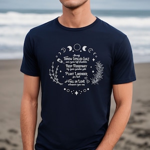 May include: Navy blue t-shirt with white text and illustrations. The text includes phrases like "Throw spilled salt" and "Plant lavender." The design features crescent moons, stars, and sprigs of herbs, creating a mystical theme.