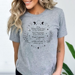 May include: Athletic heather gray t-shirt with a circular design featuring crescent moons, stars, and botanical illustrations. The text reads "Always throw spilled salt over your left shoulder, keep rosemary by your garden gate, plant lavender for luck, fall in love whenever you can."