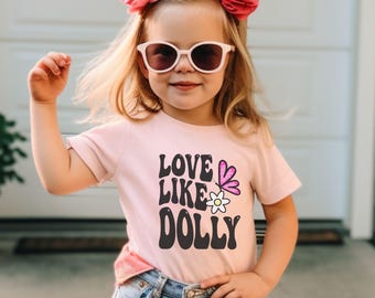 May include: A young child wearing a light pink t-shirt with the words "LOVE LIKE DOLLY" in black, along with a butterfly and flower graphic. The child is also wearing sunglasses and has pink flower hair accessories.