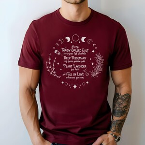 May include: A maroon t-shirt with white text and botanical illustrations. The text reads "Always throw spilled salt over your left shoulder, keep rosemary by your garden gate, plant lavender for luck, fall in love wherever you can."