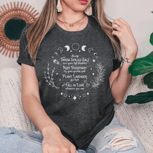 May include: Dark grey heather t-shirt with white text. The text includes phrases like "Throw spilled salt", "Keep rosemary", and "Plant lavender". The shirt is part of a spiritual or witchy theme.