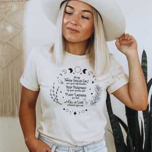 May include: Cream-colored t-shirt with a circular graphic featuring crescent moons, herbs, and text. The text includes phrases like "Throw spilled salt," "Keep rosemary," and "Plant lavender." The shirt is worn by a person wearing a white hat.