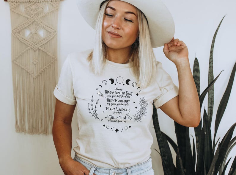 May include: Cream-colored t-shirt with a circular graphic featuring crescent moons, herbs, and text. The text includes phrases like "Throw spilled salt," "Keep rosemary," and "Plant lavender." The shirt is worn by a person wearing a white hat.