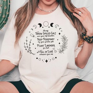 May include: Cream-colored t-shirt with a circular design featuring text and botanical illustrations. The text includes phrases like "Throw Spilled Salt" and "Plant Lavender." The word "NATURAL" is printed on the right side.