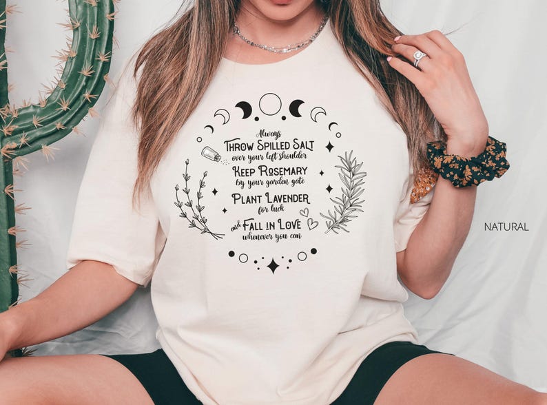 May include: Cream-colored t-shirt with a circular design featuring text and botanical illustrations. The text includes phrases like "Throw Spilled Salt" and "Plant Lavender." The word "NATURAL" is printed on the right side.