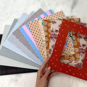 May include: A collection of rectangular fabric swatches arranged in a fan shape, featuring various patterns including gingham, stripes, polka dots, and toile. The top layer is a red fabric frame with a pink star pattern and a rectangular cutout.