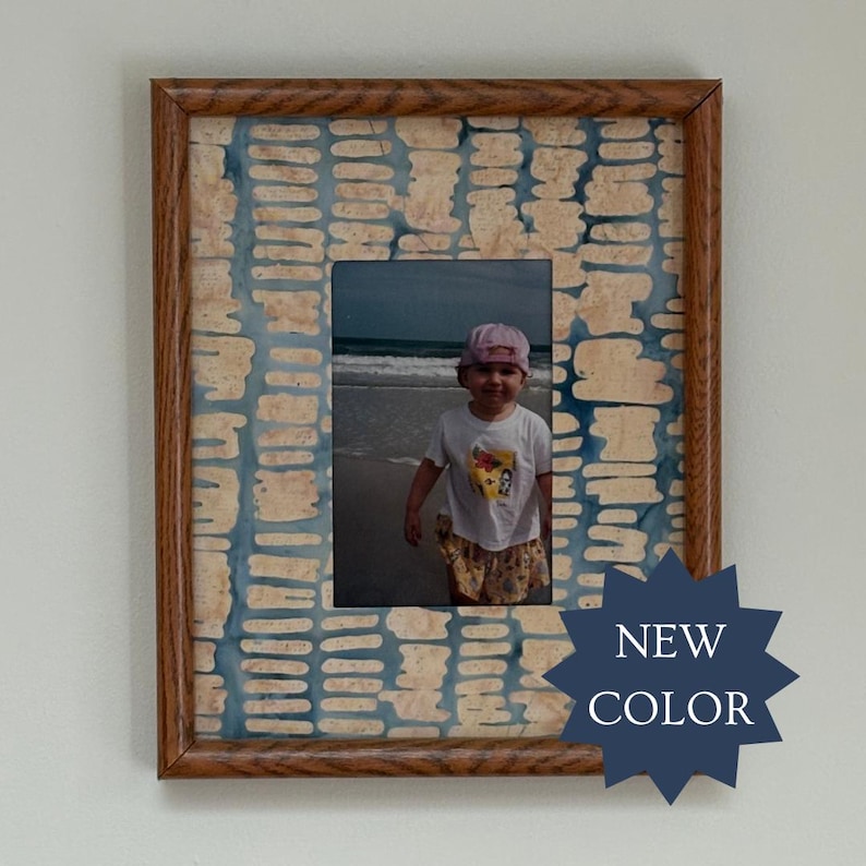 May include: A rectangular wooden picture frame featuring a decorative mat with a blue and beige abstract pattern of horizontal dashes. The frame contains a photograph of a child on a beach. A dark blue star-shaped badge displays the text NEW COLOR.