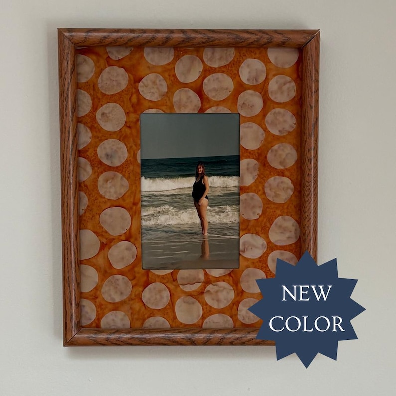 May include: A rectangular wooden picture frame featuring a border with a repeating pattern of light beige circular shapes on a vibrant orange background. The frame holds a photograph of a person standing in the ocean. A dark blue star-shaped graphic with the text NEW COLOR is overlaid on the bottom right corner.