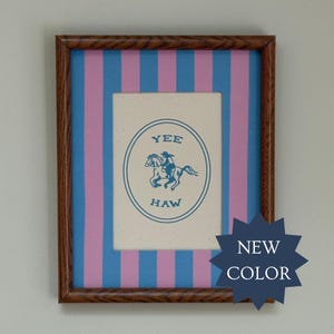 May include: A framed print featuring a cream-colored center with a blue oval border, a blue illustration of a person riding a horse, and the text YEE HAW. The background consists of vertical blue and pink stripes, all enclosed in a dark wood frame.